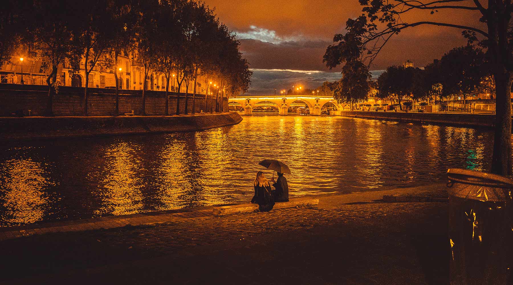 couple seated together at edge of seine river Paris at golden hour riverbank lamps lit